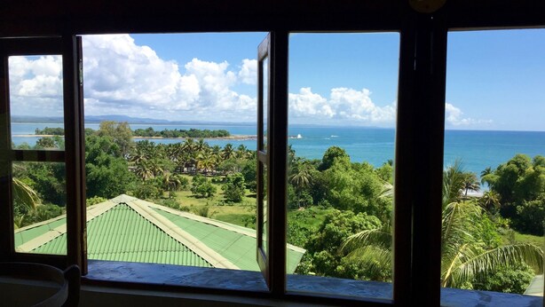 A room with a view of a beach, palm trees, and a tennis court. - Oasis Lodge (Fenoarivo Atsinanana)