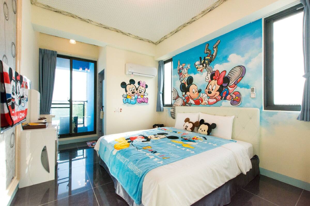 Una habitación de hotel con una colcha temática de Mickey Mouse, una lavadora y un ventanal con cortinas.