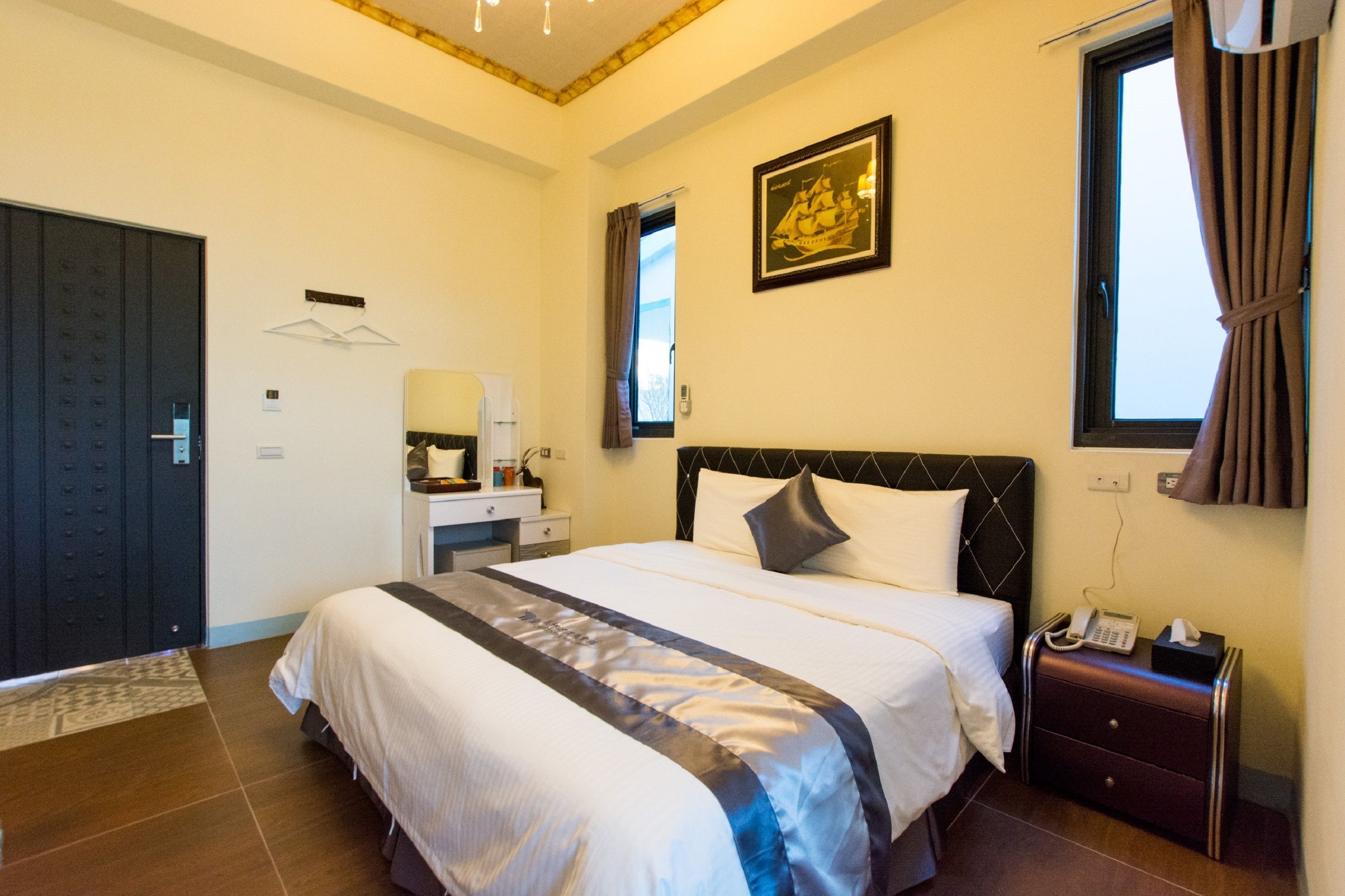 A hotel room with a large bed, a bedside table with a lamp, a window with curtains, a door, and a wall-mounted phone.