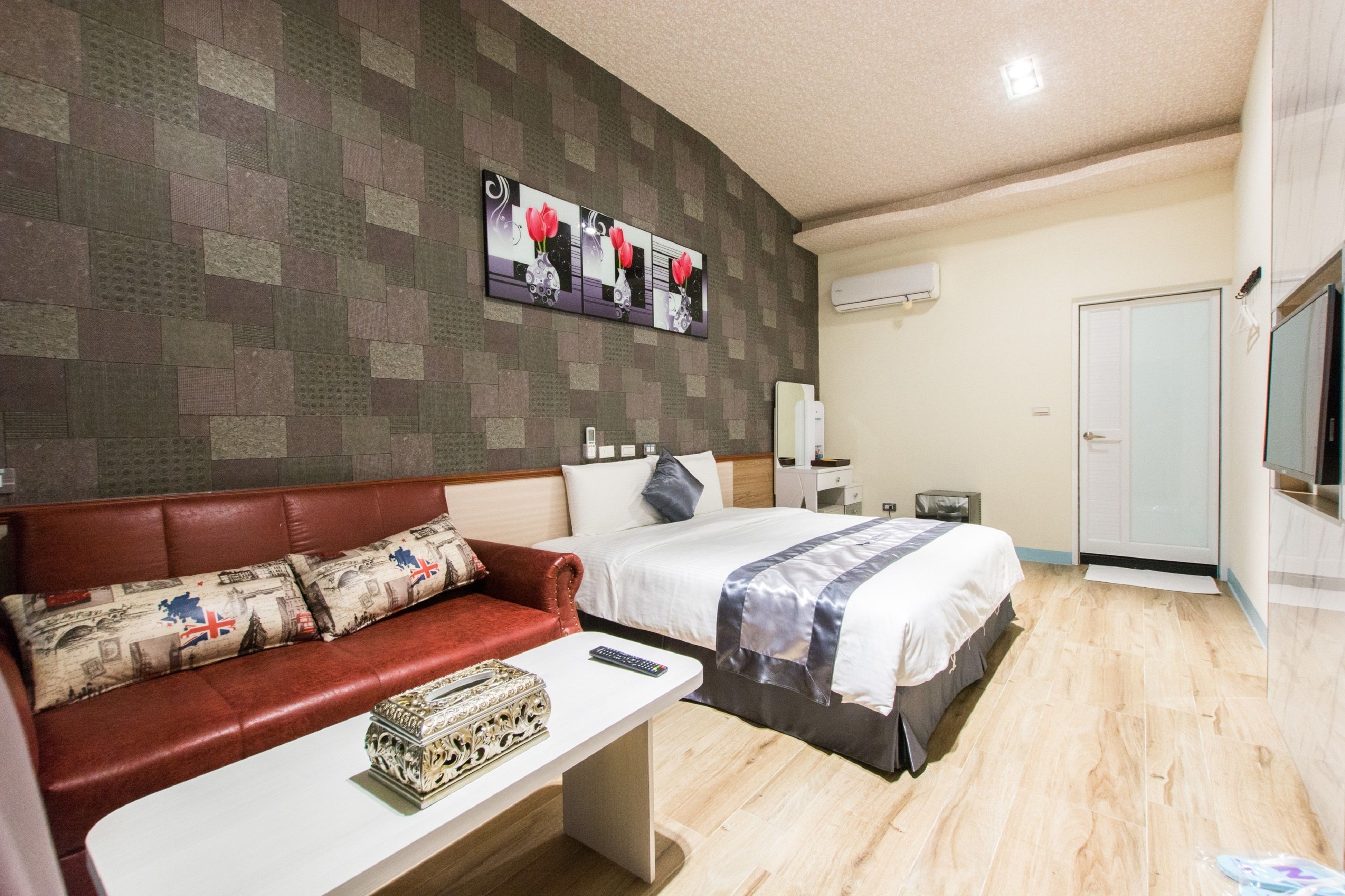 Una habitación de hotel con un sofá de cuero rojo, una cama con edredón gris y blanco, una mesita con un control remoto y un televisor montado en la pared.