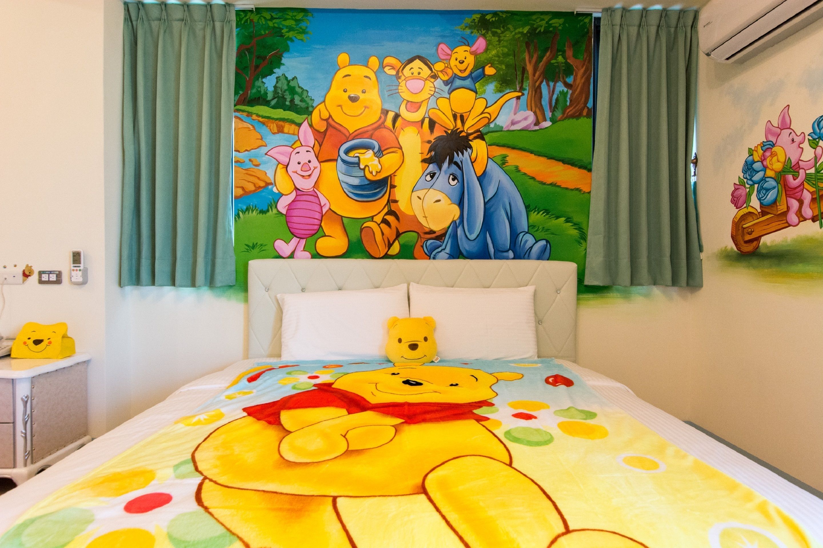 Une chambre d’enfant avec une couverture de lit aux motifs de Winnie l’ourson, une fresque représentant les personnages de Winnie l’ourson sur le mur, et une petite table de chevet sur laquelle est posé un ours en peluche.
