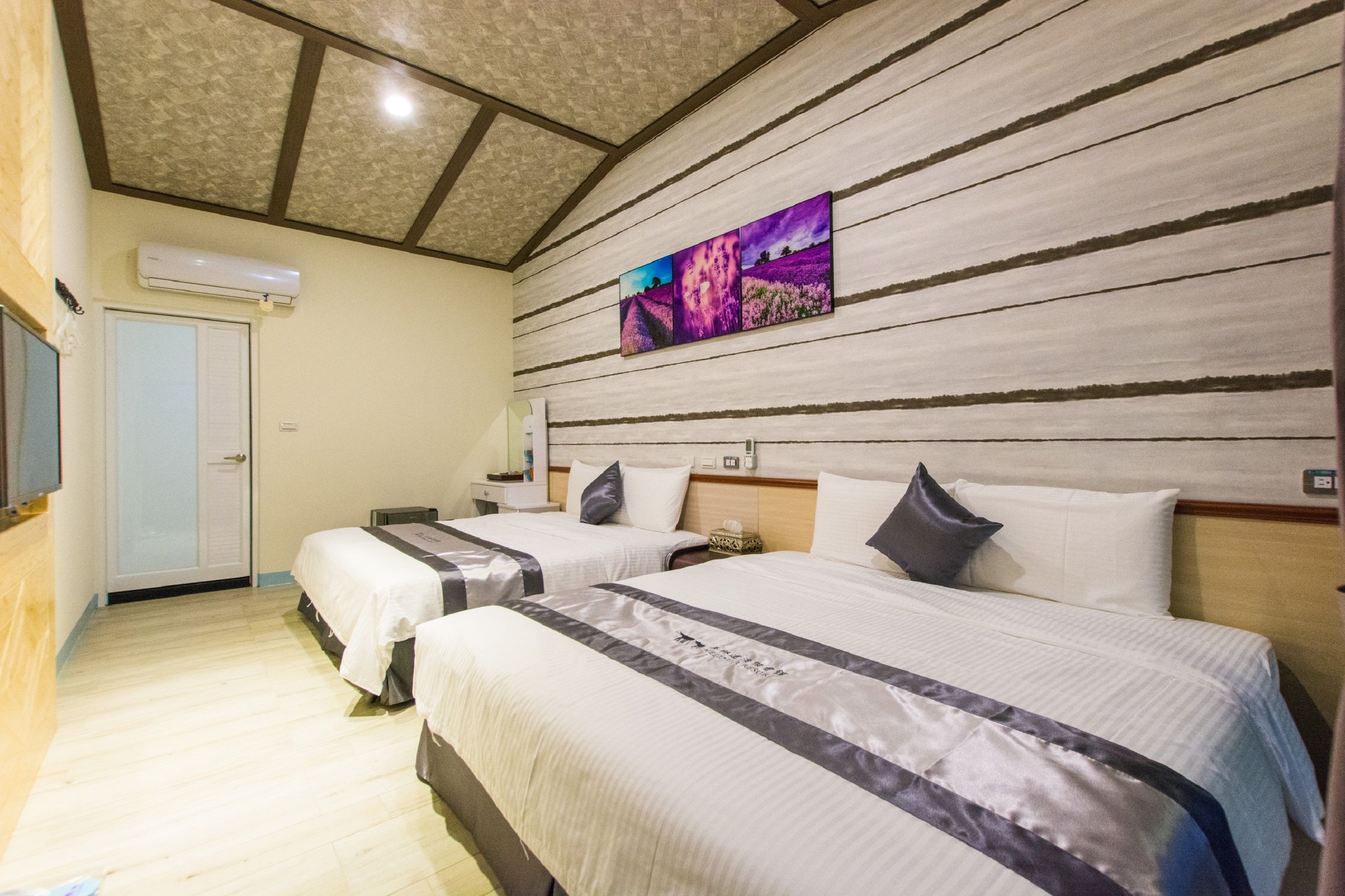 A hotel room with two beds, a wooden headboard, a ceiling with a wooden beam design, and a wall-mounted air conditioner.