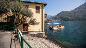Un edificio con balcón que da a un lago, una lancha amarrada cerca y montañas al fondo.