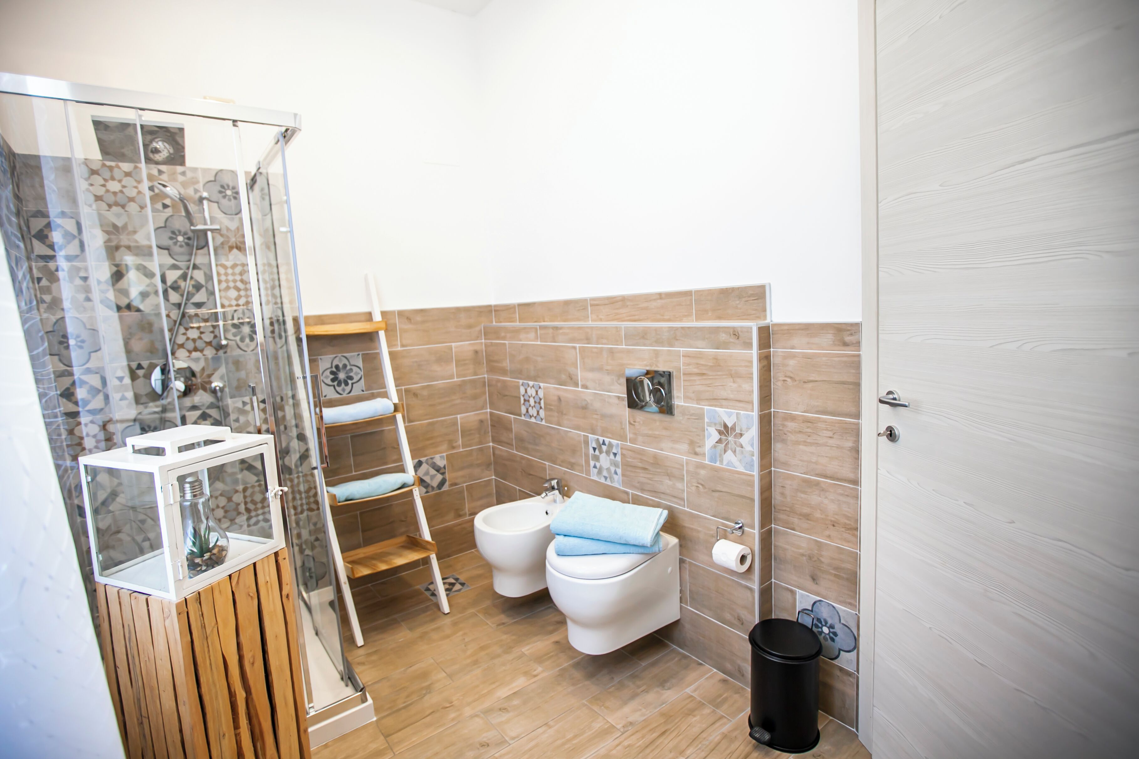 A bathroom with a shower enclosure, two toilets, a towel rack, a trash bin, and a wooden shelf with a decorative item.