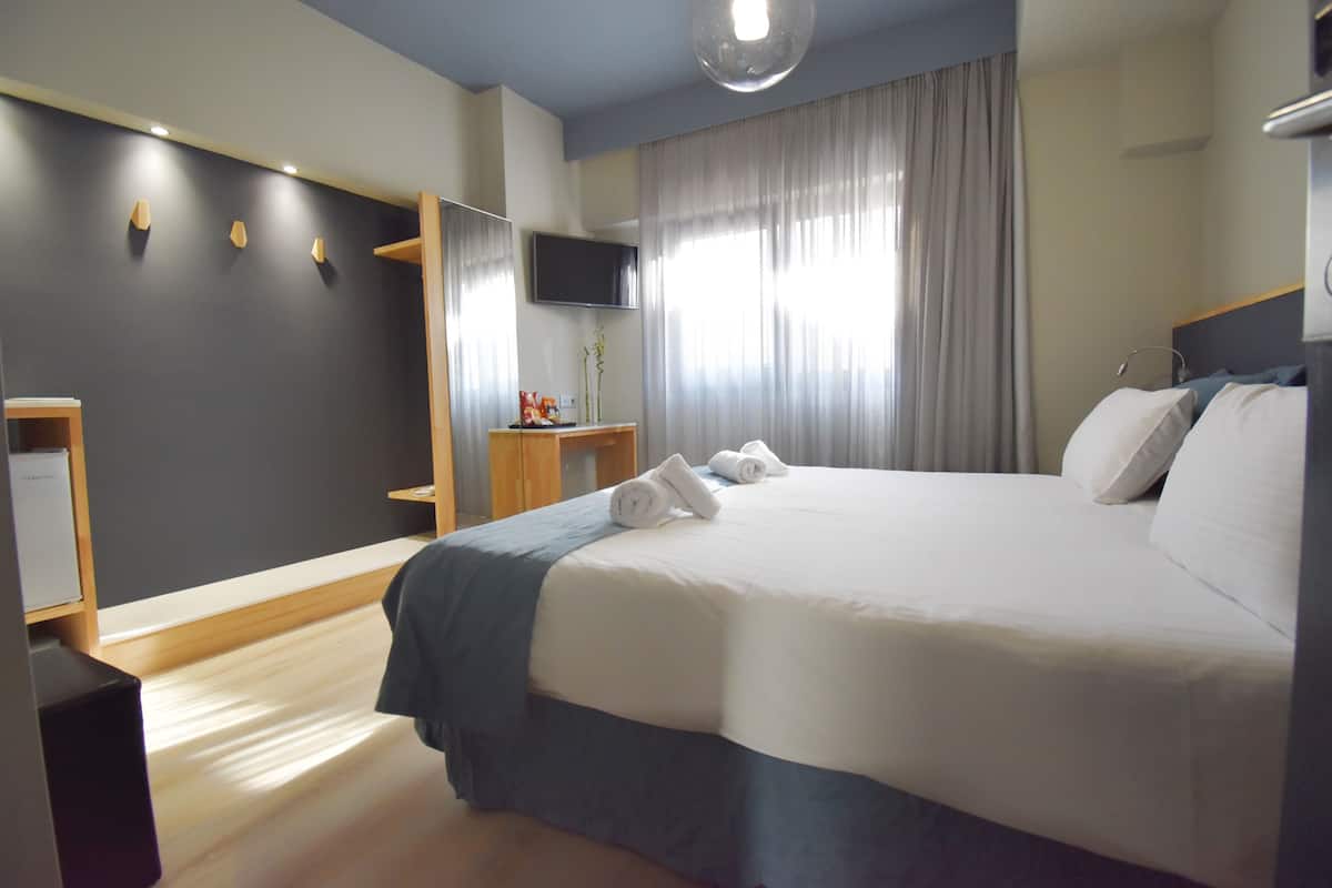 A modern hotel room with a large bed, a flat-screen TV, a wooden nightstand, and a large window with curtains.