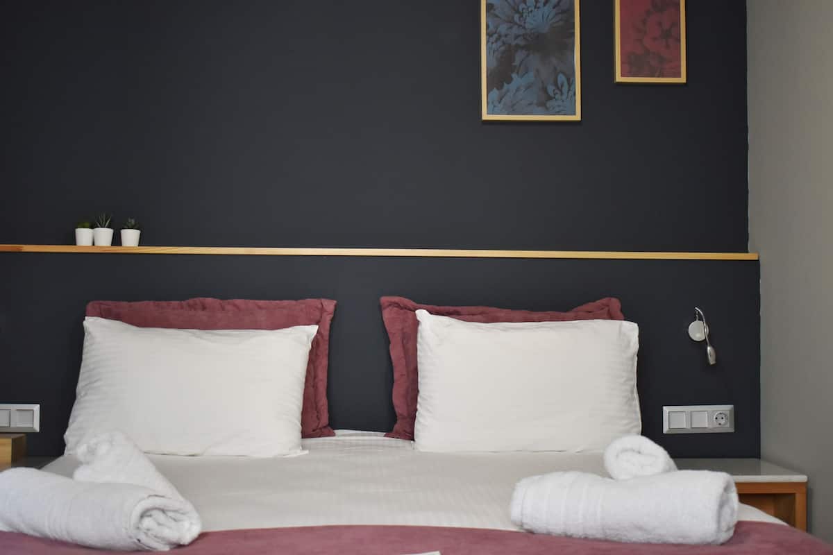 A hotel room with a bed, two pillows, a bedside table, a nightstand, a wall-mounted phone, and two small potted plants.