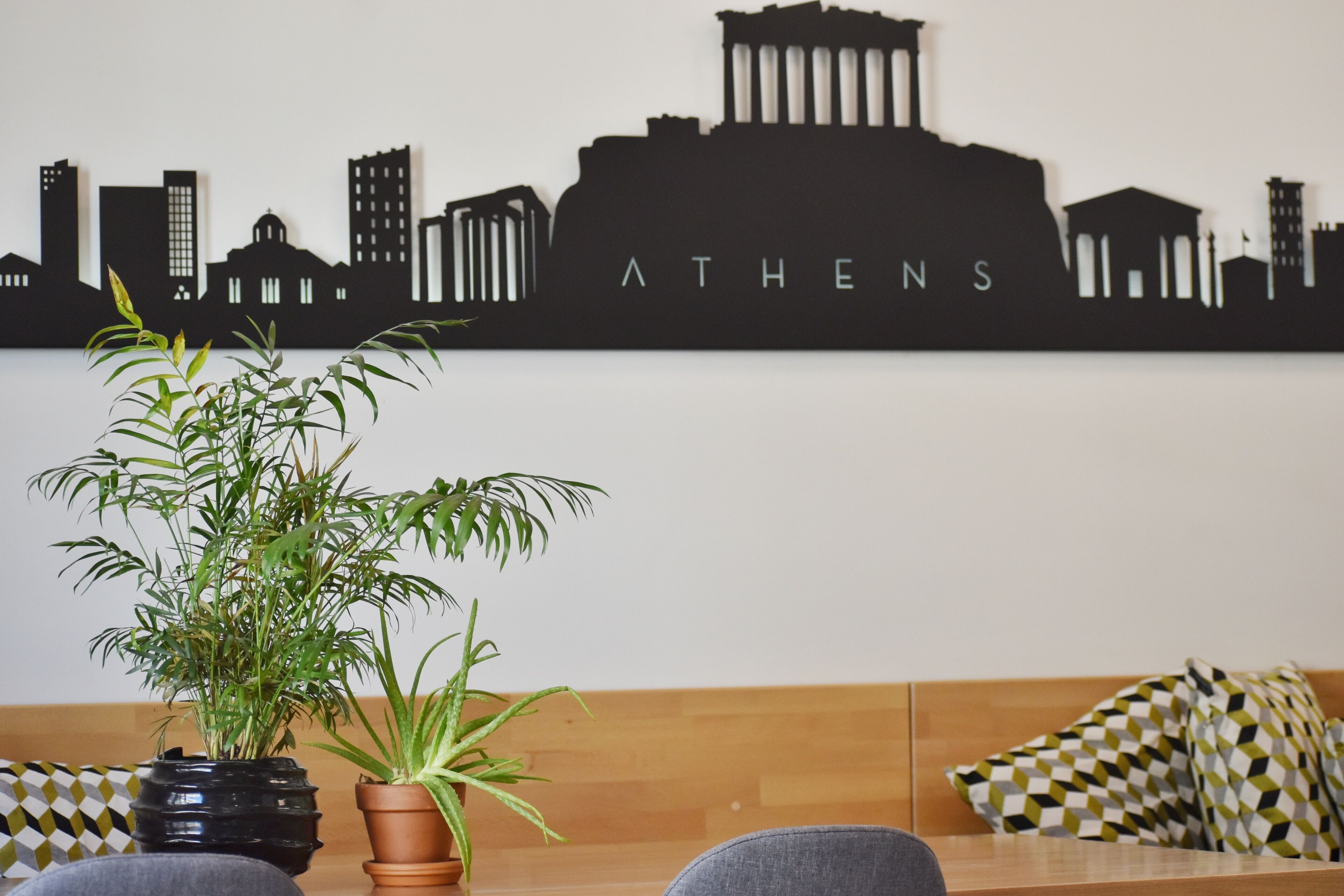 A room with a wooden bench, a potted plant, and a wall with a black silhouette of a city skyline labeled "ATHENS".