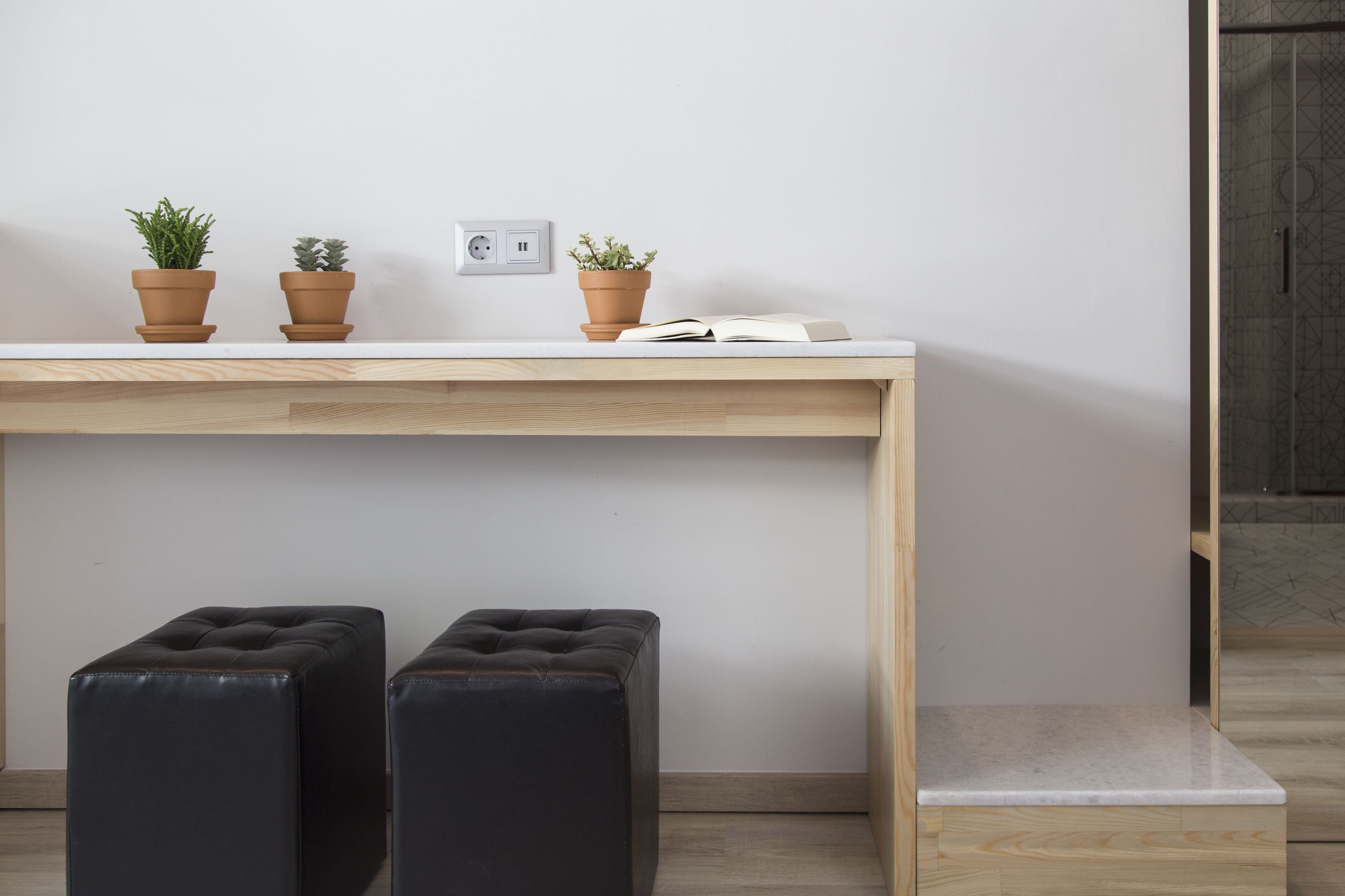 A minimalist room with a wooden desk, two potted plants, a book, and two black ottomans.