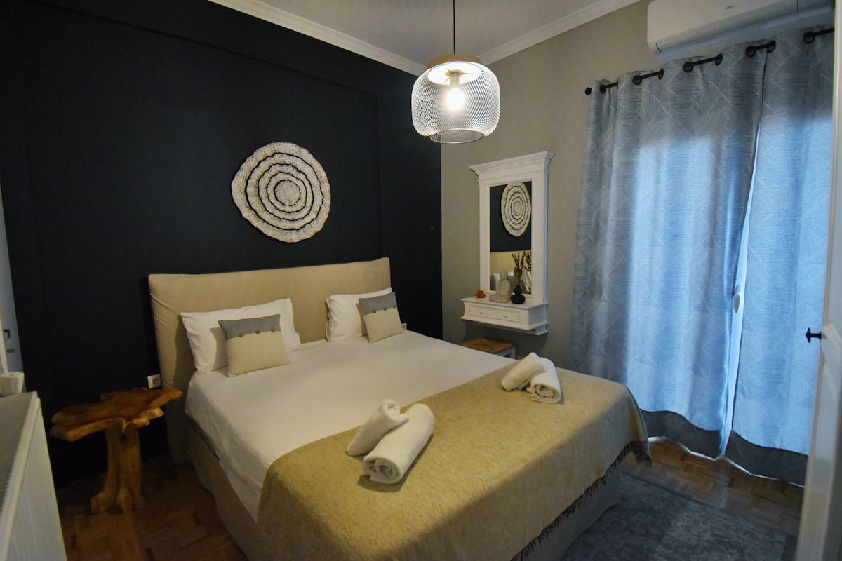 A neatly made bed with a beige blanket and two rolled towels, a round wall decoration, a hanging light fixture, a mirror above a small table, and a blue curtain.