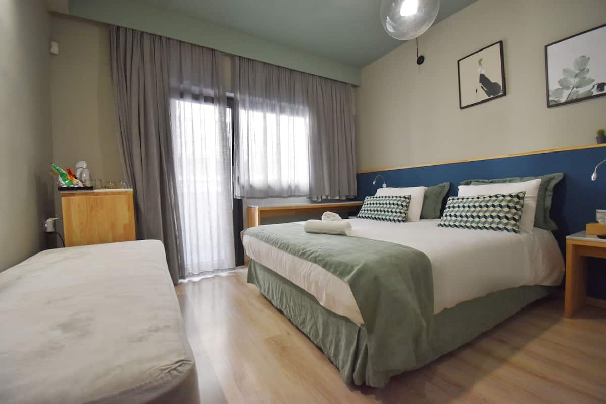 A hotel room with a large bed, a smaller bed, a wooden nightstand, a bedside table, a window with curtains, and wall art.