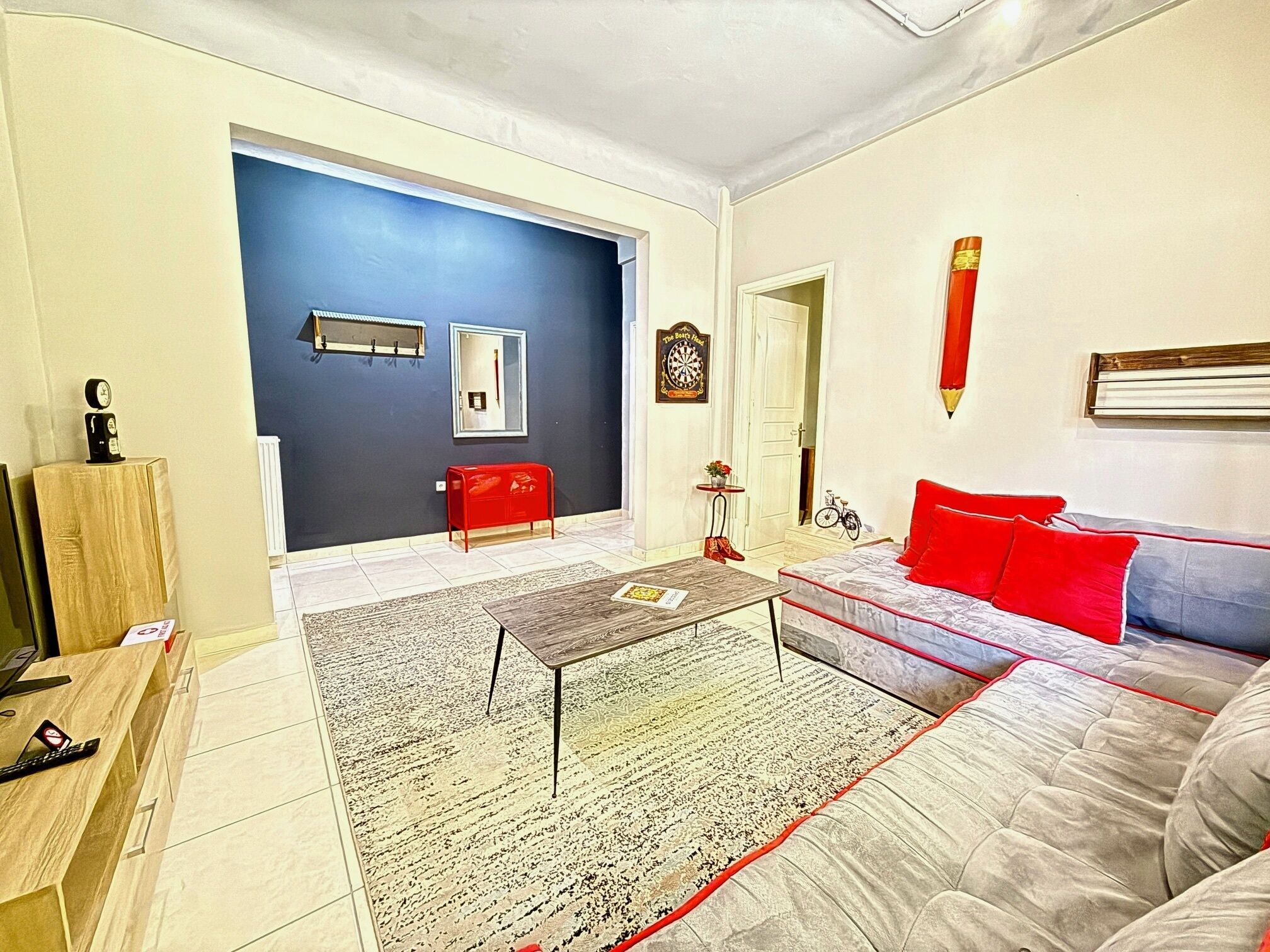 Una sala de estar con un sofá, una mesa de centro y un banco rojo. Hay un espejo en la pared y un reloj encima.