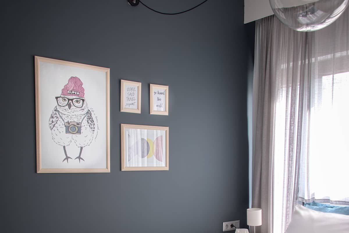 A modern hotel room with a bed, a bedside table with a hat and a magazine, a wall with framed artwork, and a window with curtains.