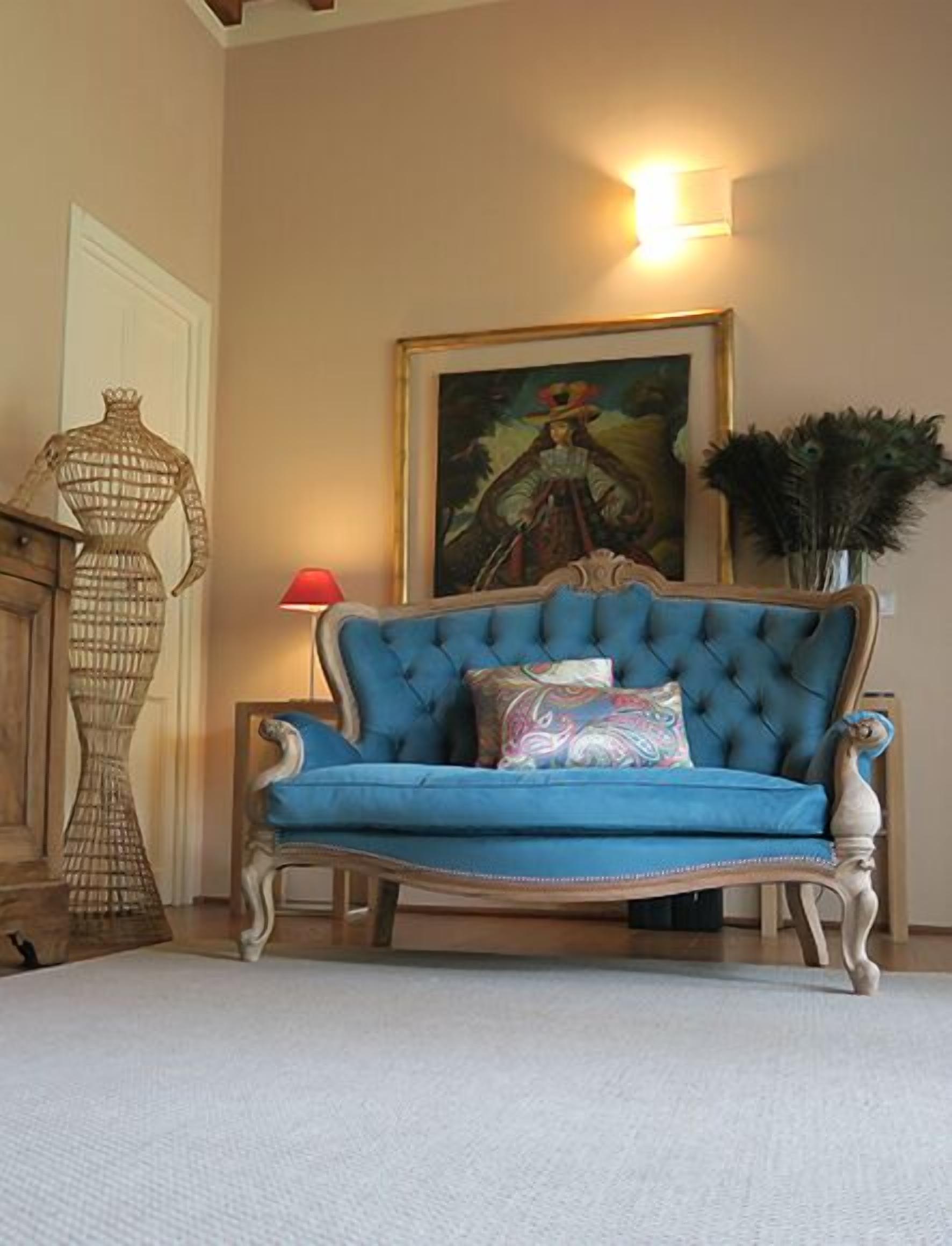 A blue upholstered sofa with a tufted backrest and decorative pillows, positioned in a room with a beige wall and a framed painting above it.