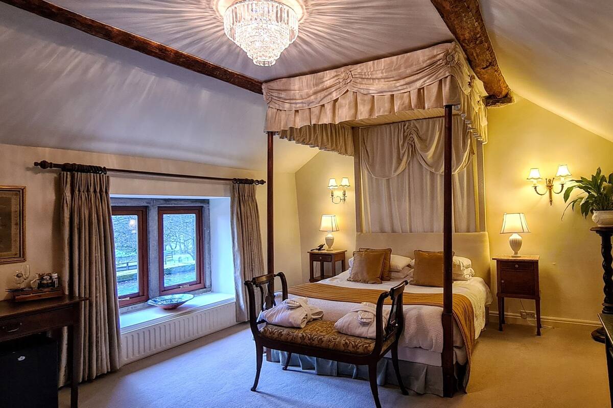 A four-poster bed with a canopy, a desk with a kettle, a chair, and a window with curtains.