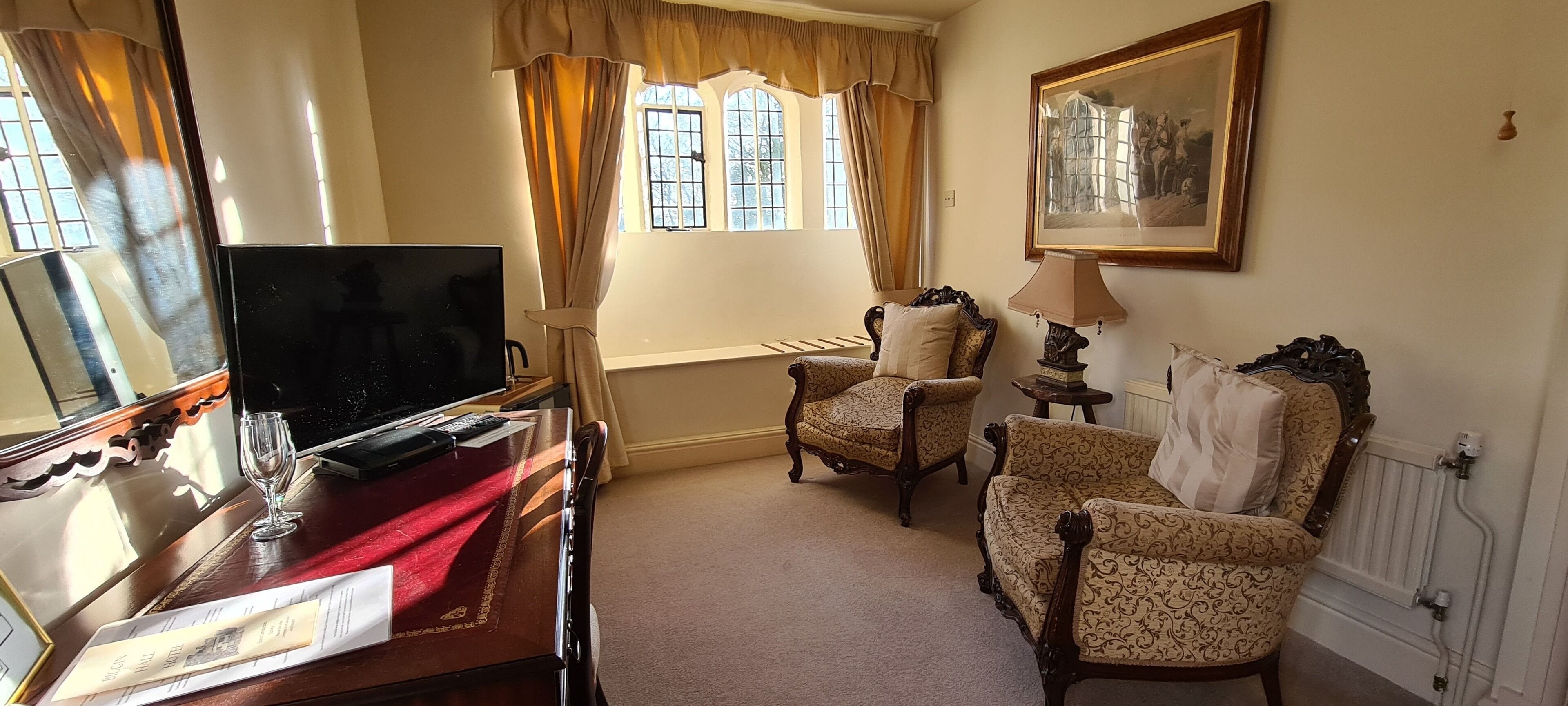 A room with a TV, a desk with a glass, a lamp, and two patterned chairs.