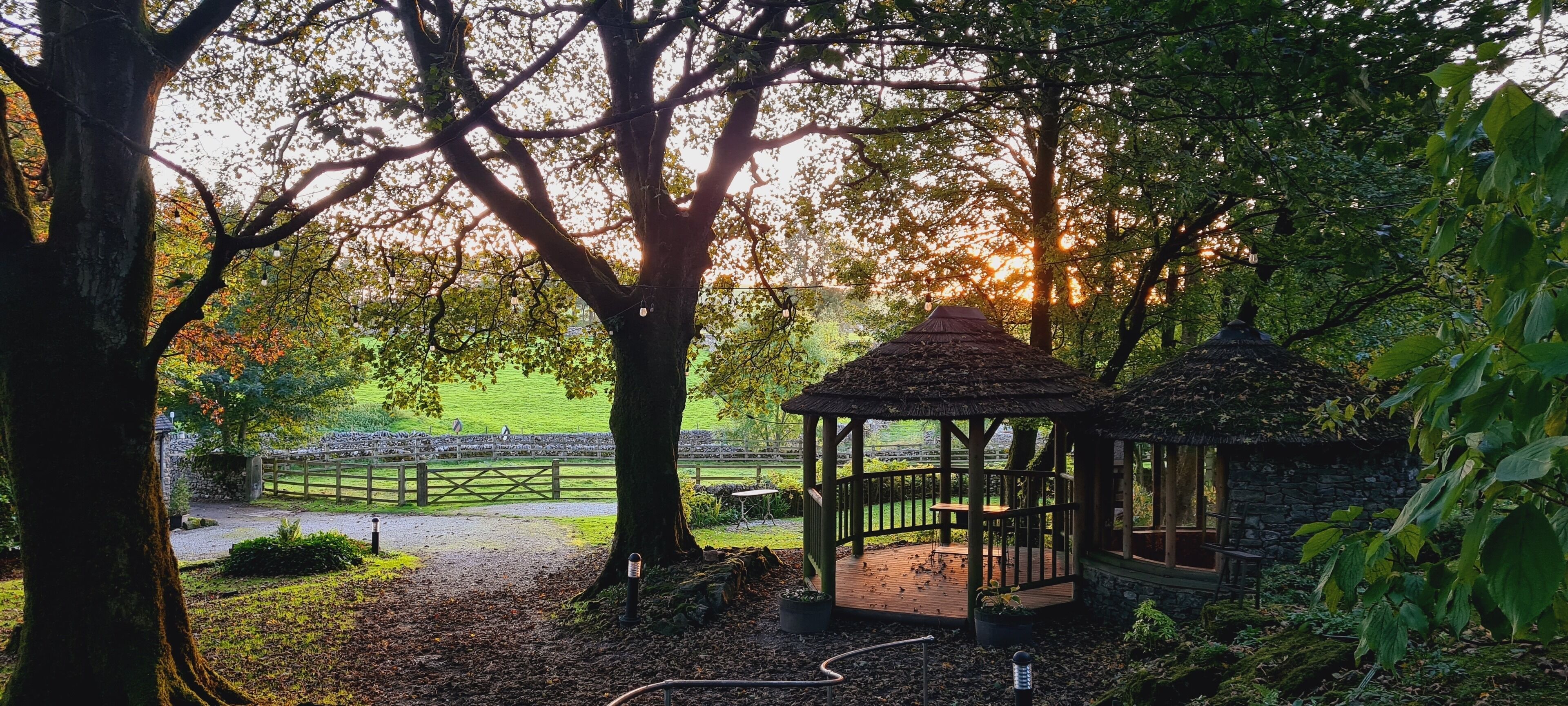 A gazebo with a thatched roof surrounded by trees and a stone wall.