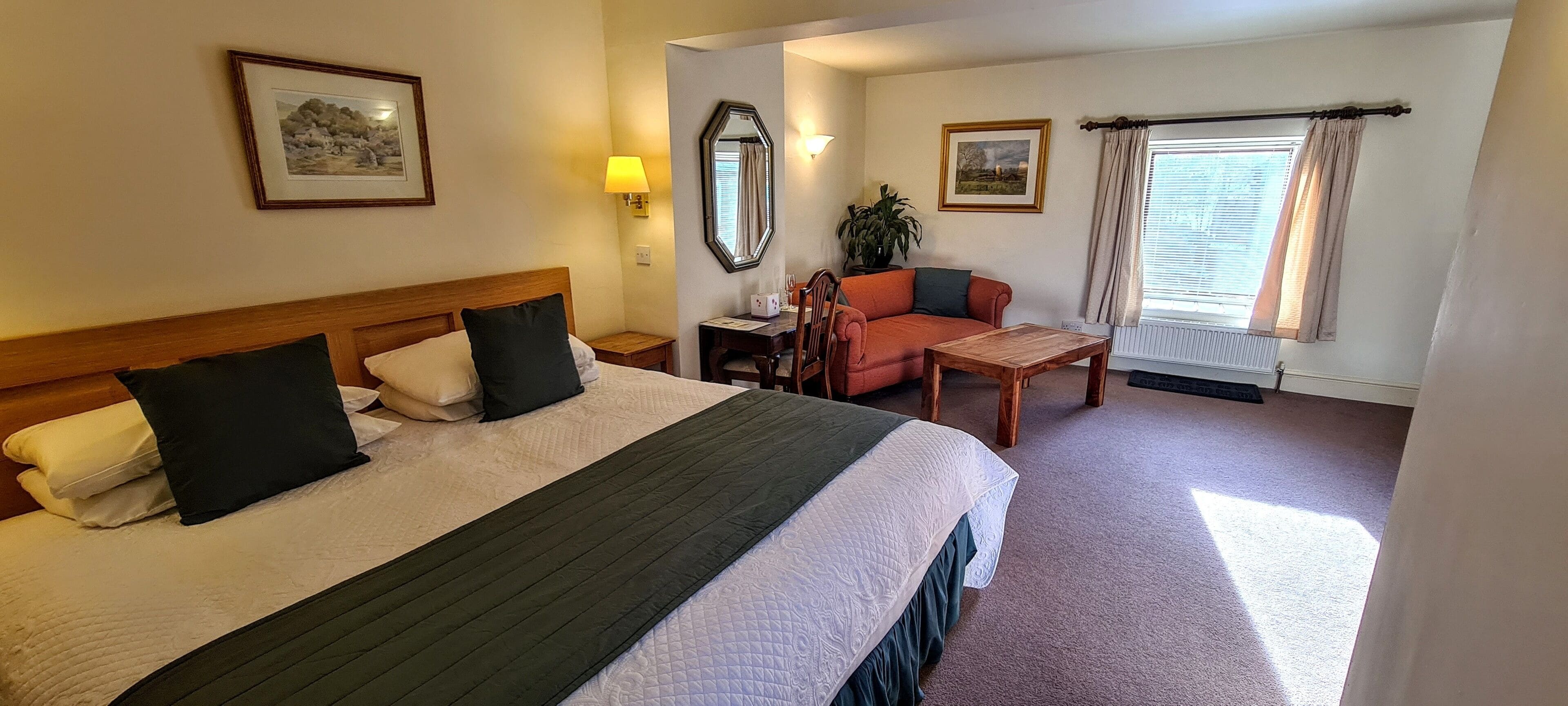 Habitación de hotel con una cama grande, cabecera de madera, escritorio con silla, una mesita y dos cuadros en las paredes.