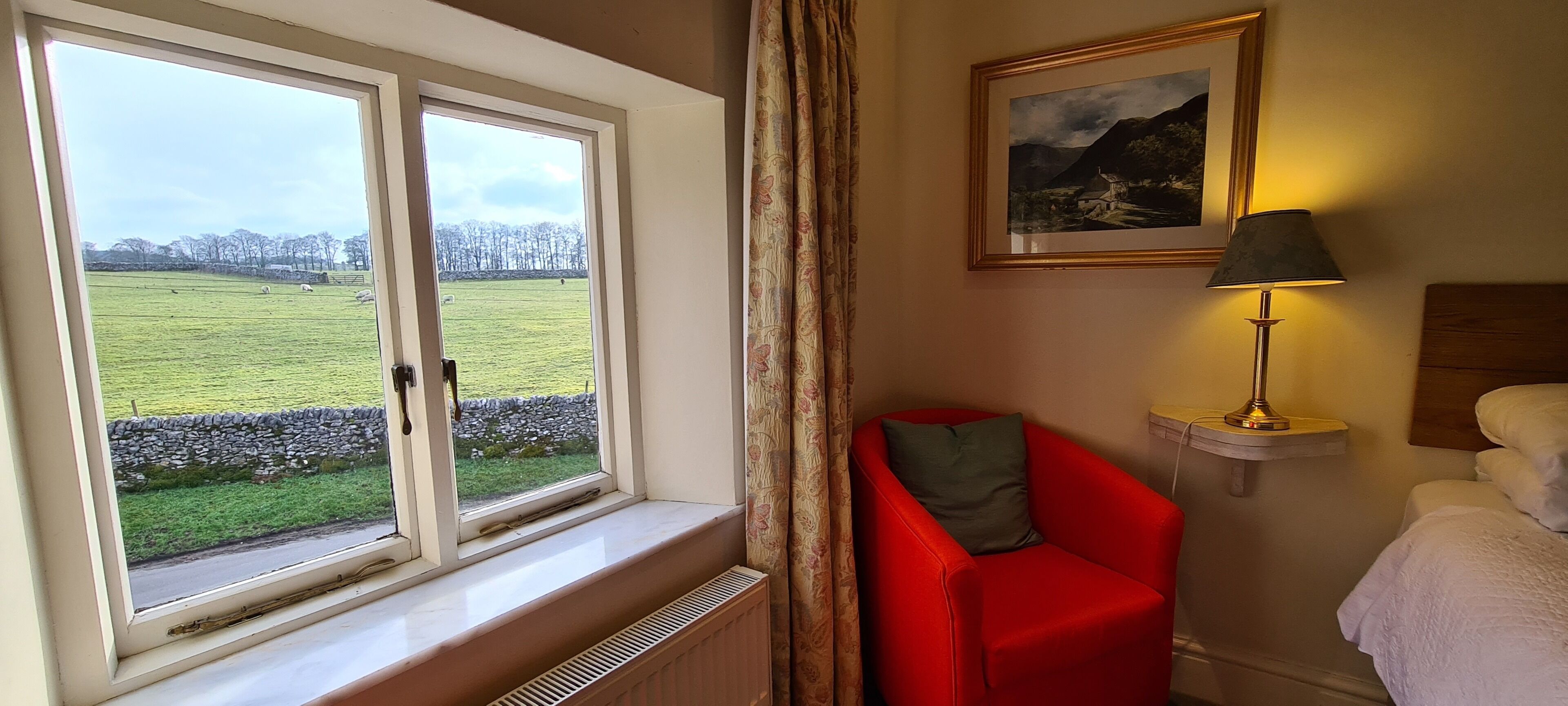 Una habitación con un gran ventanal que da a un campo verde, una silla roja, una mesita de noche con una lámpara y una cama con ropa blanca.