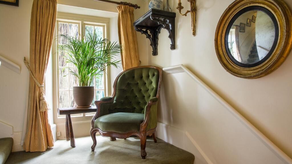 Un fauteuil vert recouvert de tissu, dans une pièce avec une fenêtre, une plante en pot, un miroir et une petite table d’appoint en bois foncé.