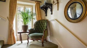 Un fauteuil vert recouvert de tissu, dans une pièce avec une fenêtre, une plante en pot, un miroir et une petite table d’appoint en bois foncé.