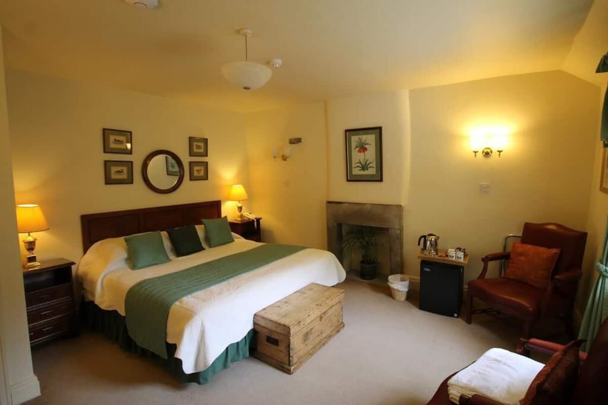 A bedroom with a large bed, bedside lamps, a fireplace, a chair, and a small table with a kettle.