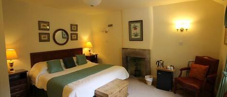 A bedroom with a large bed, bedside lamps, a fireplace, a chair, and a small table with a kettle.