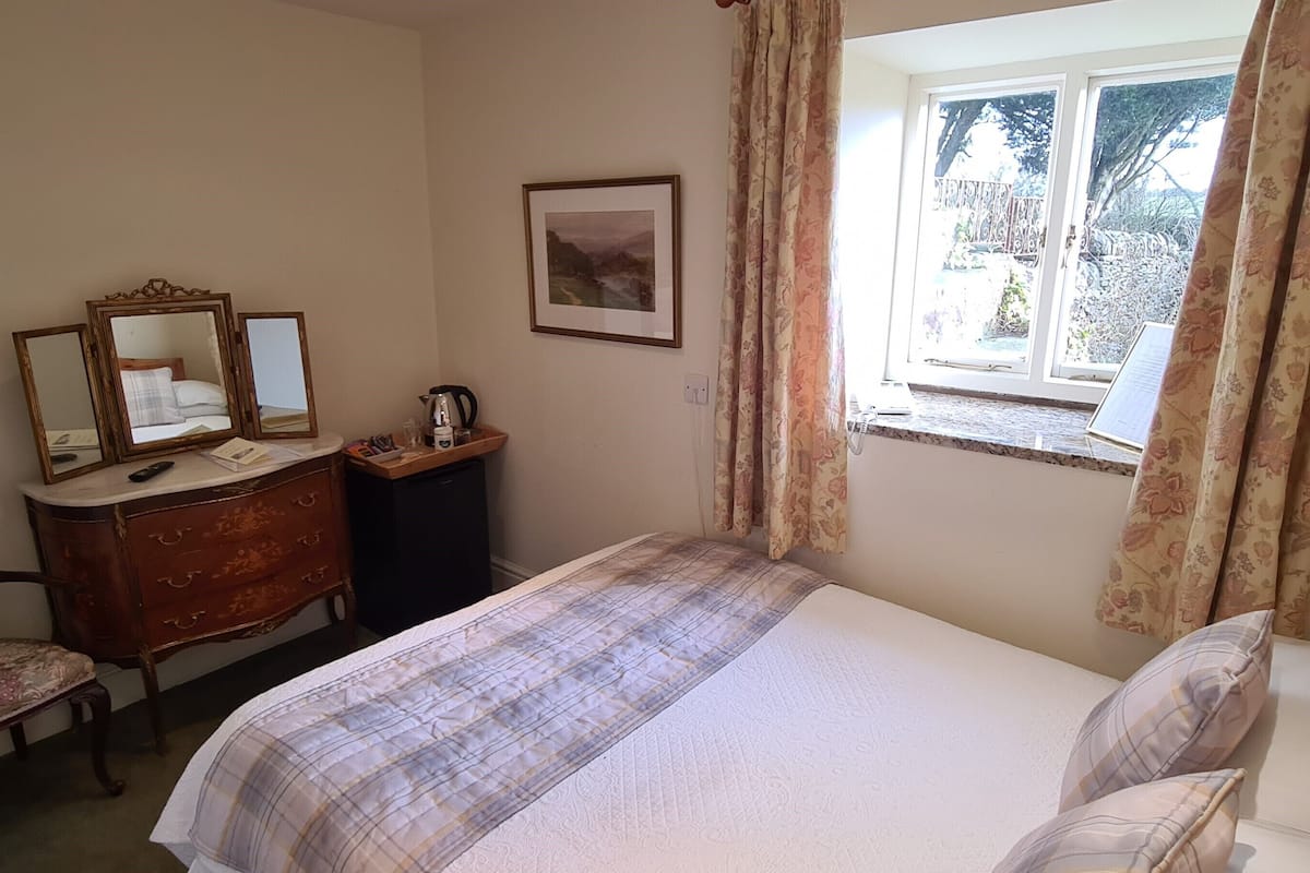 A bedroom with a bed, a dresser with mirrors, a chair, a window with curtains, and a framed picture on the wall.