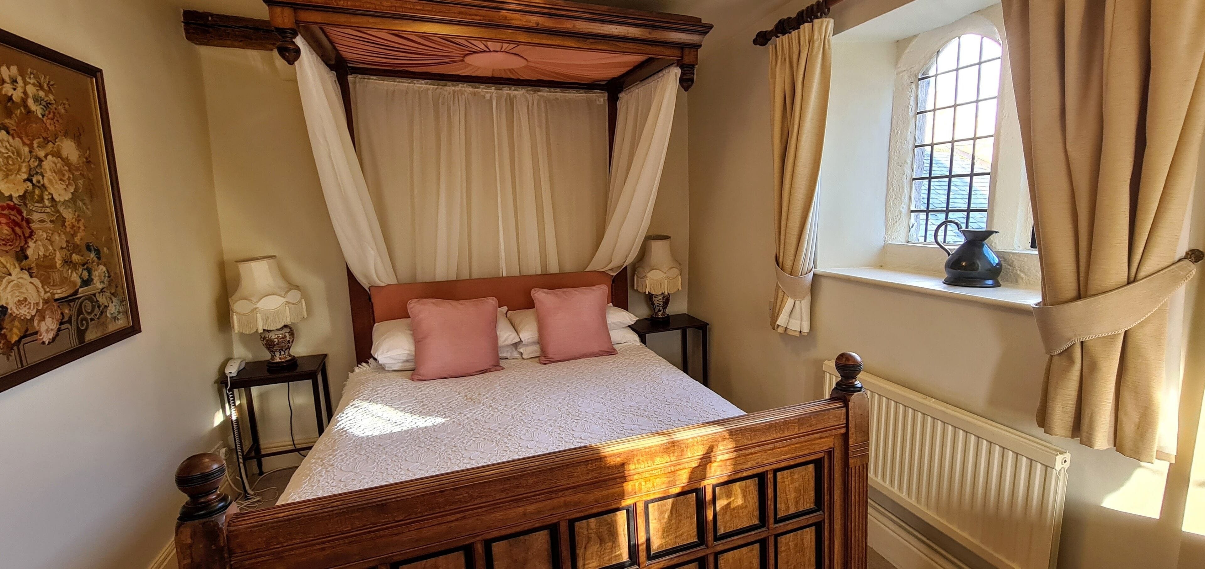 Una cama con dosel y cuatro columnas, con edredón blanco y almohadas rosas, una mesita de noche con lámpara y una ventana con cortinas.