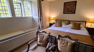 A bedroom with a large bed, two bedside tables with lamps, a radiator, a window with curtains, and a chair with cushions.