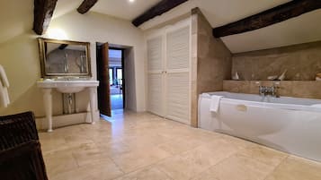A bathroom with a bathtub, a sink, and a mirror. There is a wicker chair and a door leading to another room.