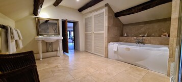 A bathroom with a bathtub, a sink, and a mirror. There is a wicker chair and a door leading to another room.