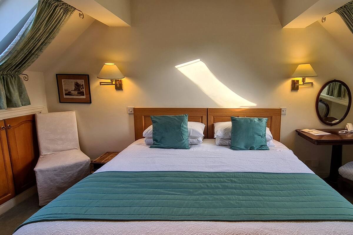 A bedroom with a large bed, two bedside lamps, a wooden nightstand, a chair, and a desk with a telephone.