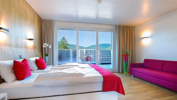 A modern hotel room with a large bed, a balcony with a view, a bright pink sofa, and a wooden wall. - Hotel Rhy (Oberriet)
