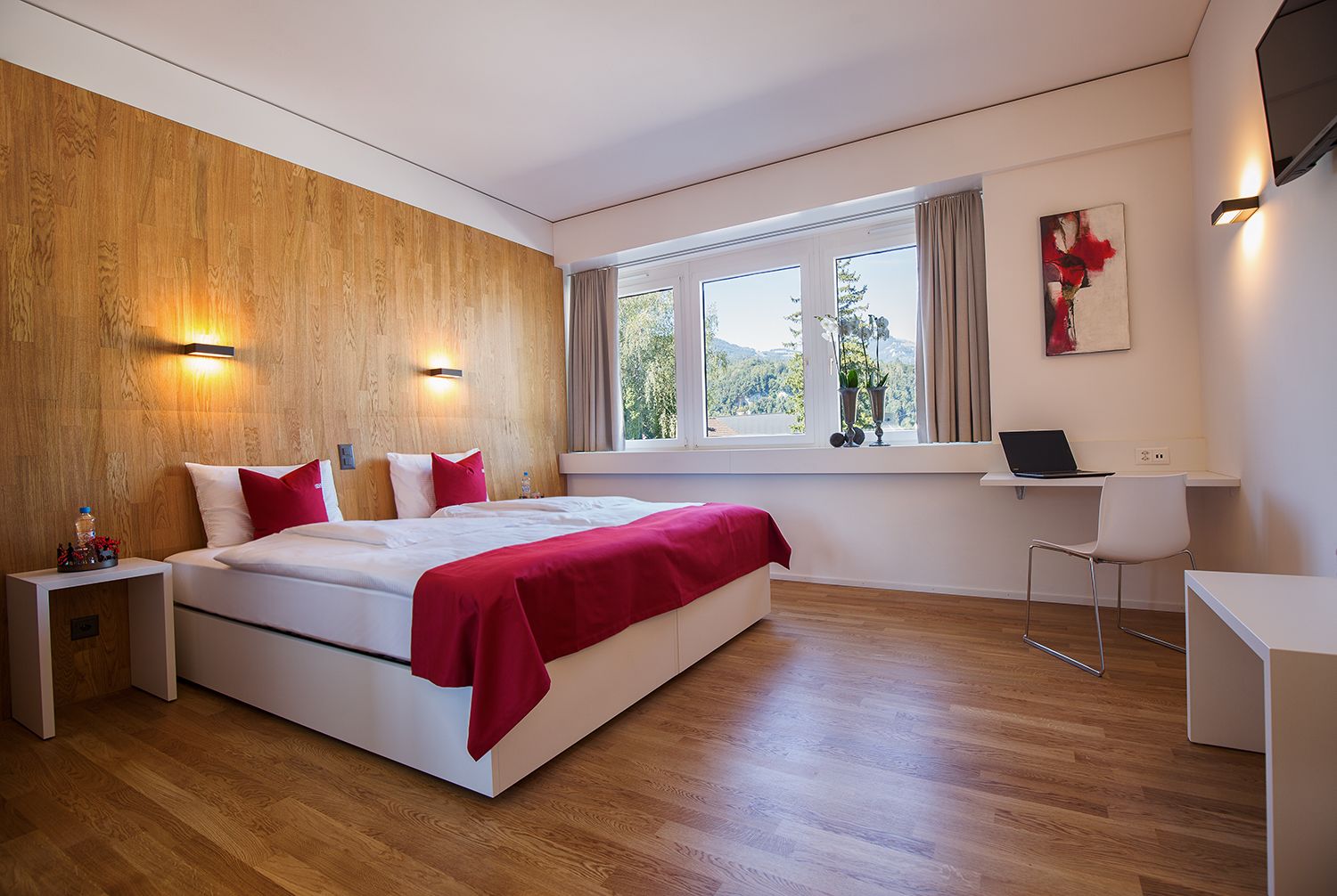 Un dormitorio moderno con una pared de acento de madera, una cama grande con ropa de cama roja, un escritorio con una computadora portátil y vistas a zonas verdes a través de la ventana.