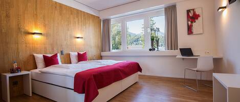 Un dormitorio moderno con una pared de acento de madera, una cama grande con ropa de cama roja, un escritorio con una computadora portátil y vistas a zonas verdes a través de la ventana.