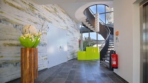 A modern lobby with a marble wall, a wooden planter with flowers, a bright green reception desk, a staircase, and a fire extinguisher. - Hotel Rhy (Oberriet)