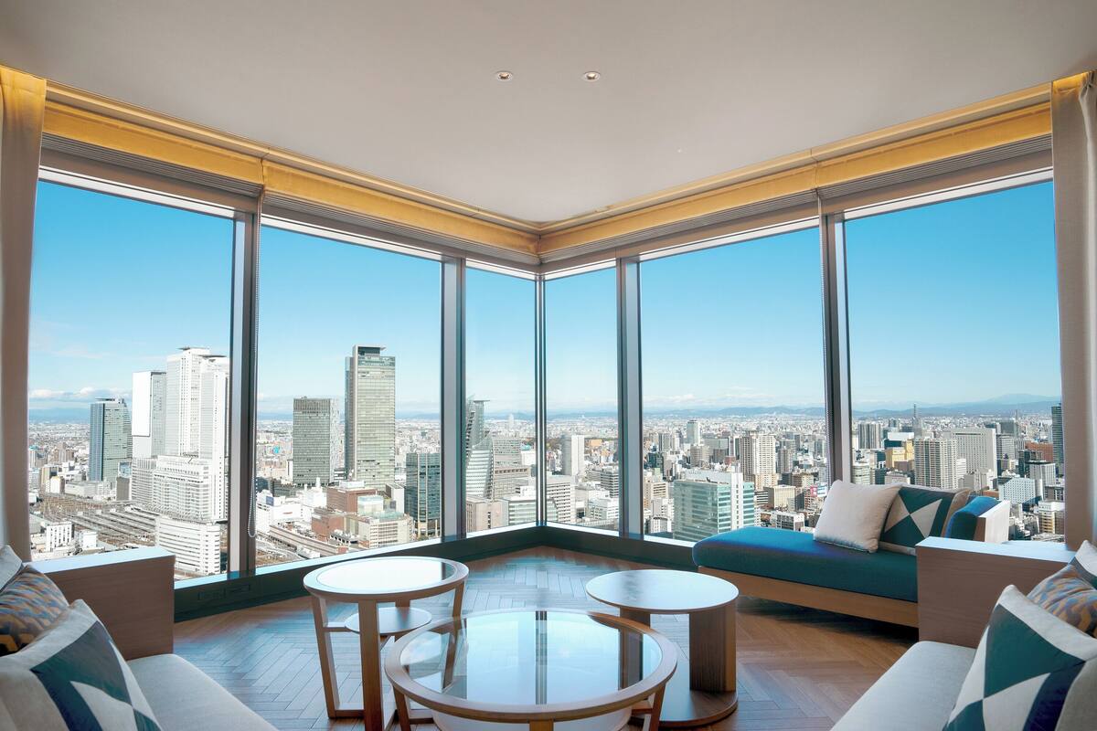 A high-rise room with large windows offering a city view, a sofa, a small table, and a chair.
