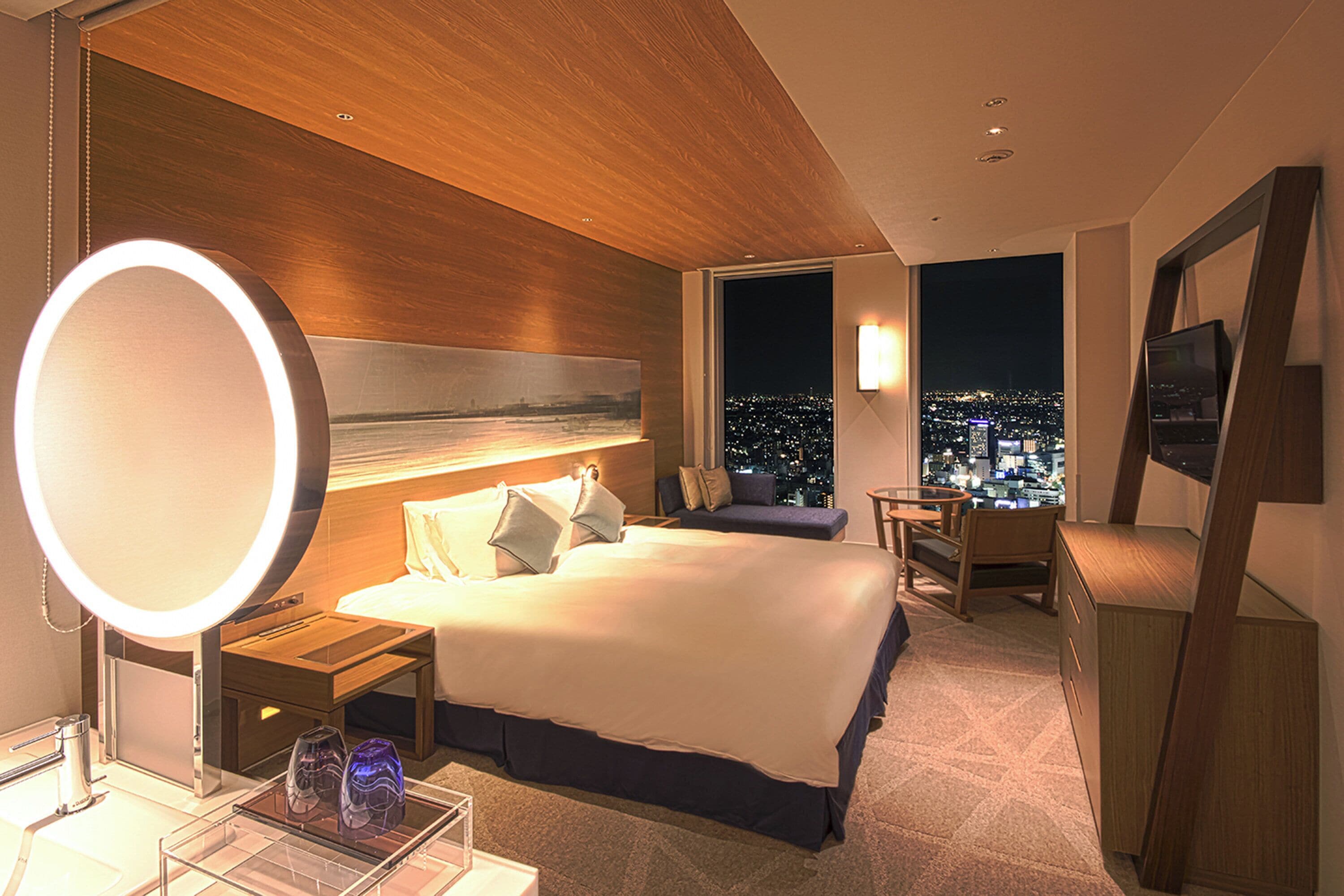 A modern hotel room with a large bed, bedside table, and a round mirror. There is a desk with a chair, a window with a city view, and a wall-mounted TV.