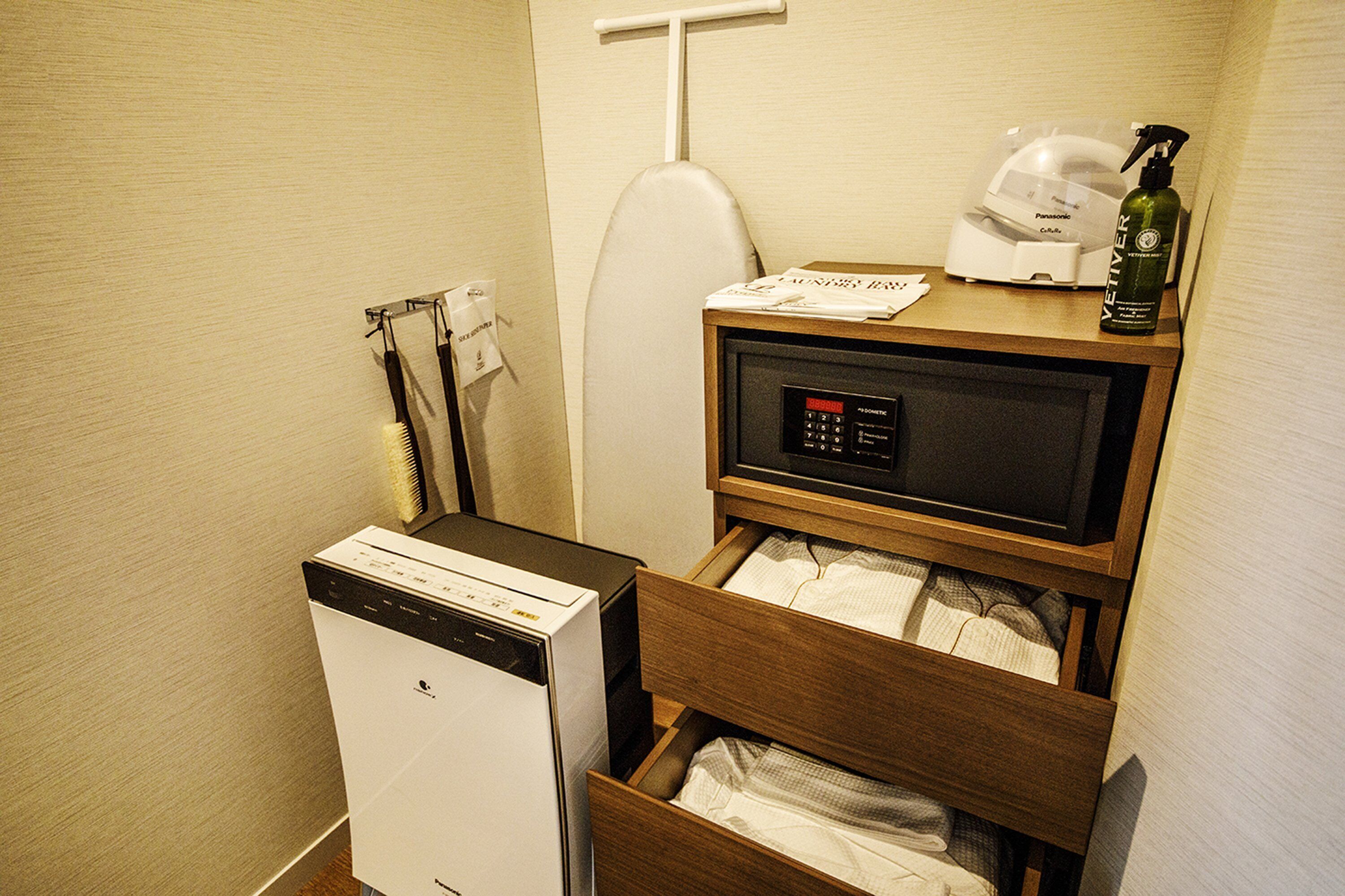 Habitació d'hotel compacta amb rentadora i assecadora integrades, microones, una petita prestatgeria de fusta amb una ampolla i un dispensador de paper higiènic.