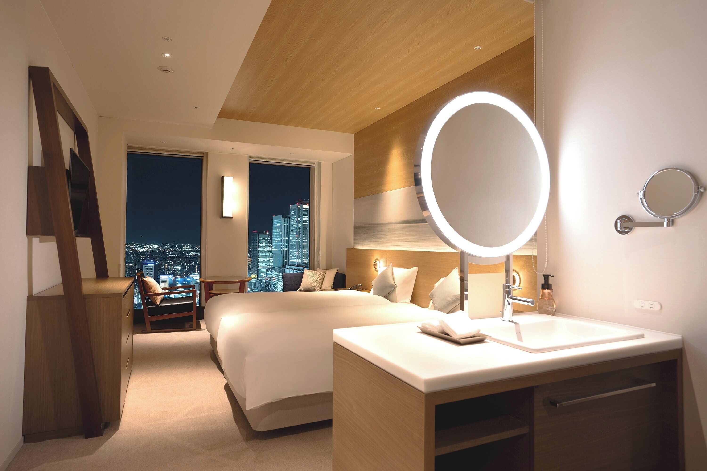 A modern hotel room with a large bed, a bedside table, a round mirror, and a view of the city at night.