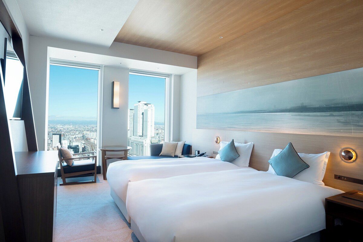 A modern hotel room with two large beds, a city view, a painting on the wall, and a desk with a chair.