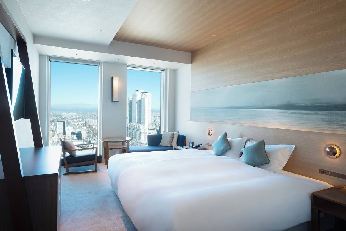 A modern hotel room with a large bed, a seating area with a view, and a cityscape visible through the windows.