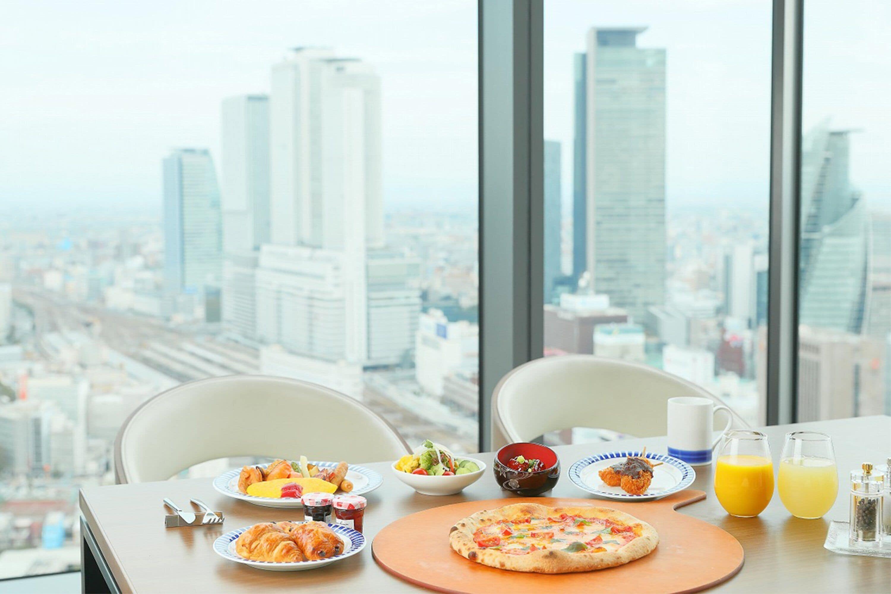 A breakfast table with plates of food, a pizza, and glasses of orange juice, set against a cityscape view.