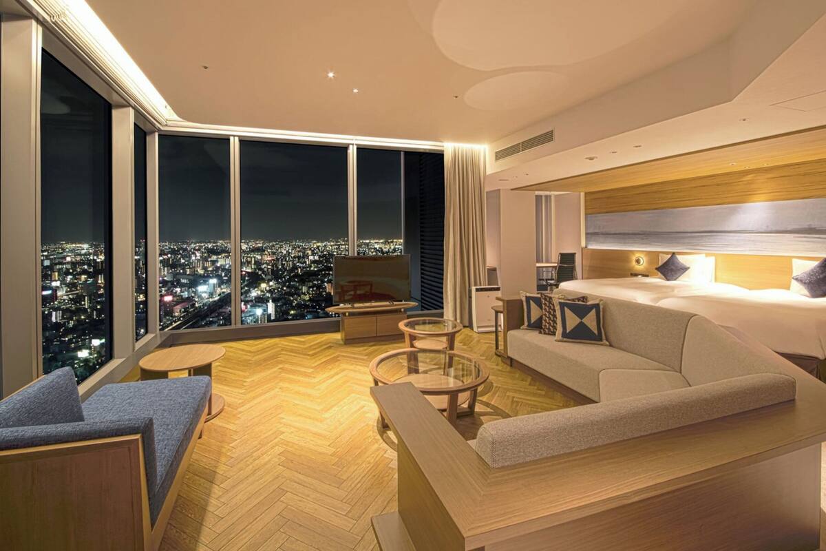 A modern hotel room with a large city view, a sofa, a bed, and a small table.