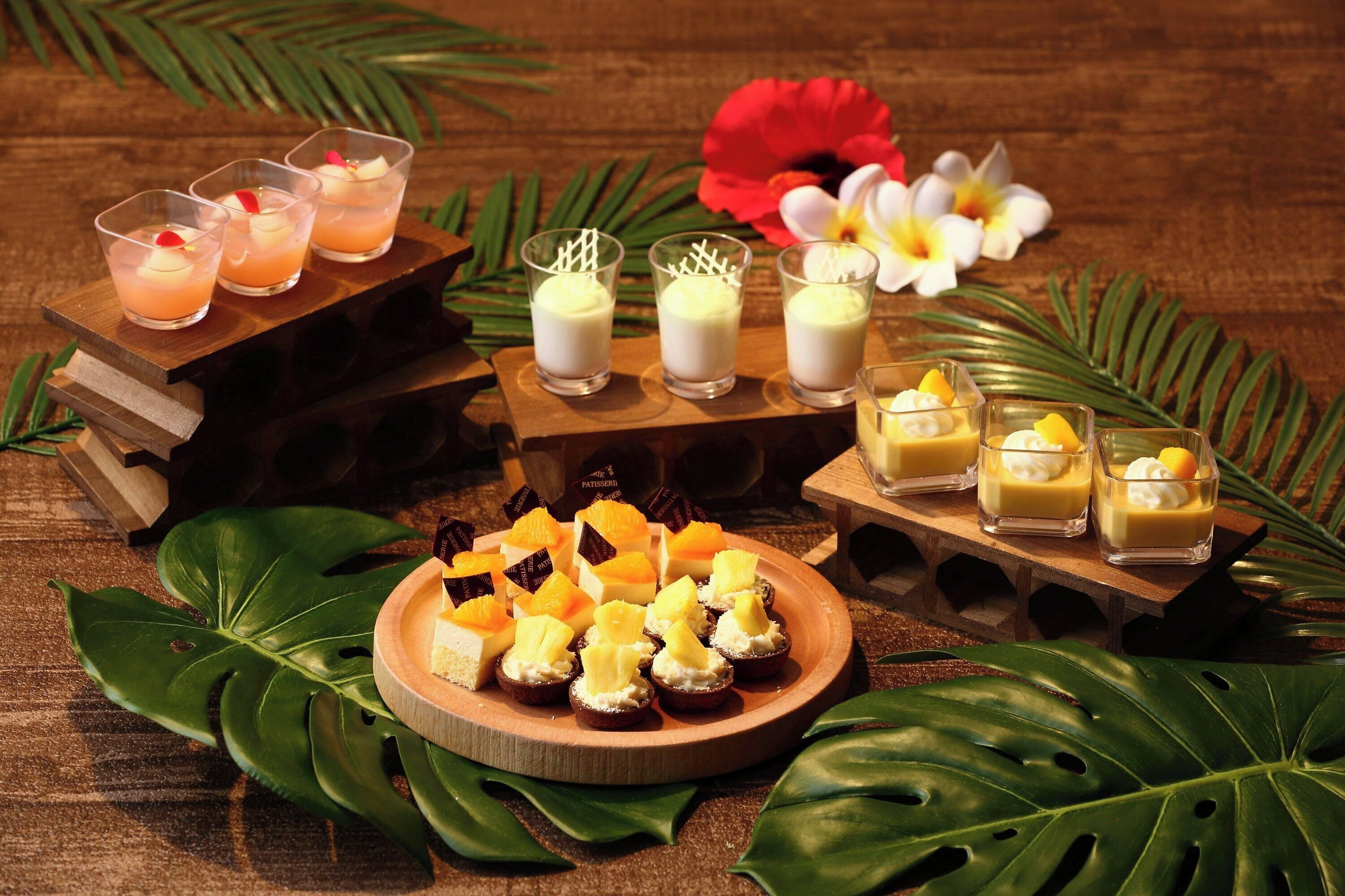 Una mesa de madera con bebidas y postres, rodeada de hojas y flores tropicales.
