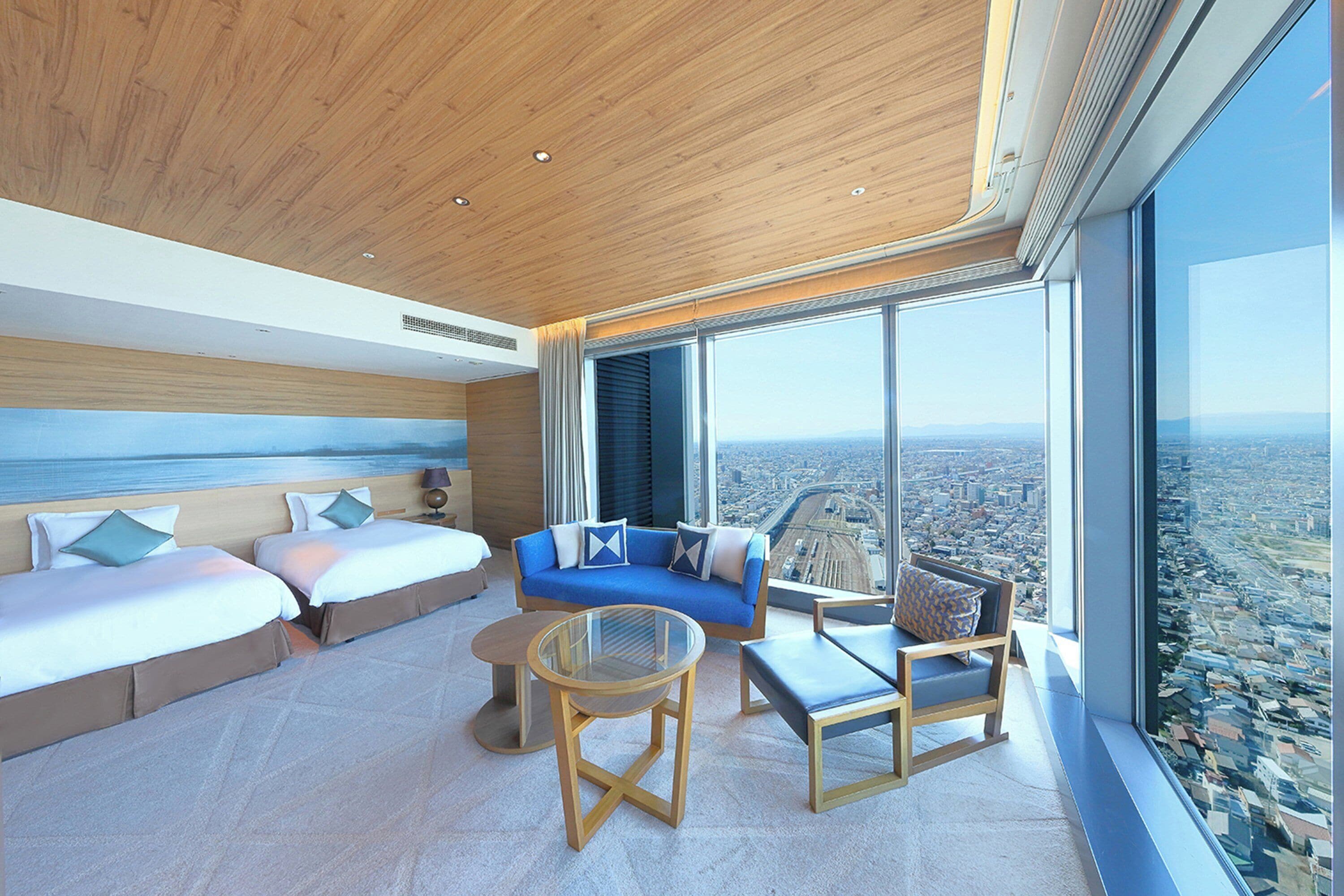 A modern hotel room with a large city view, a sofa, two beds, and a round table.