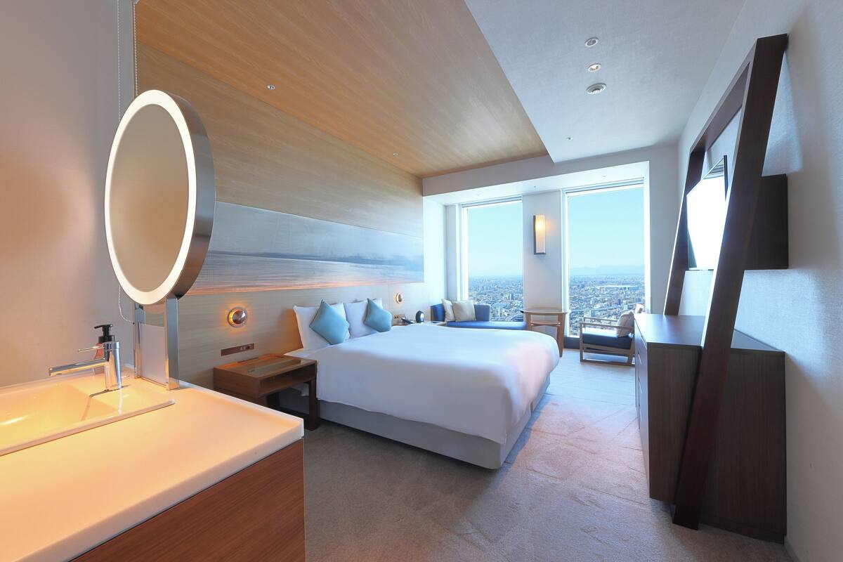 A modern hotel room with a large bed, a bedside table, a bathroom with a sink, and a view of the city.