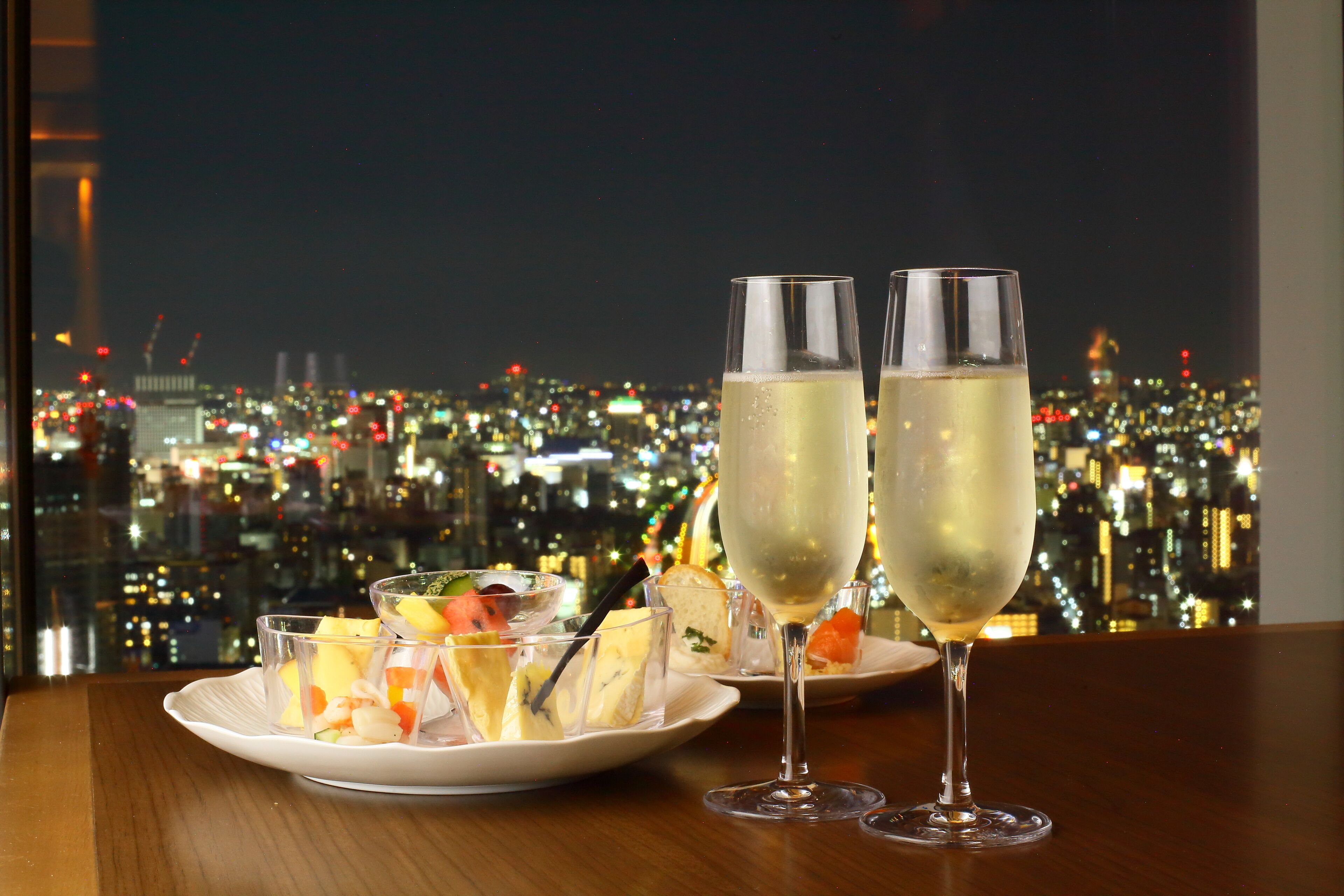 A table with two champagne flutes and a plate of appetizers against a cityscape view.