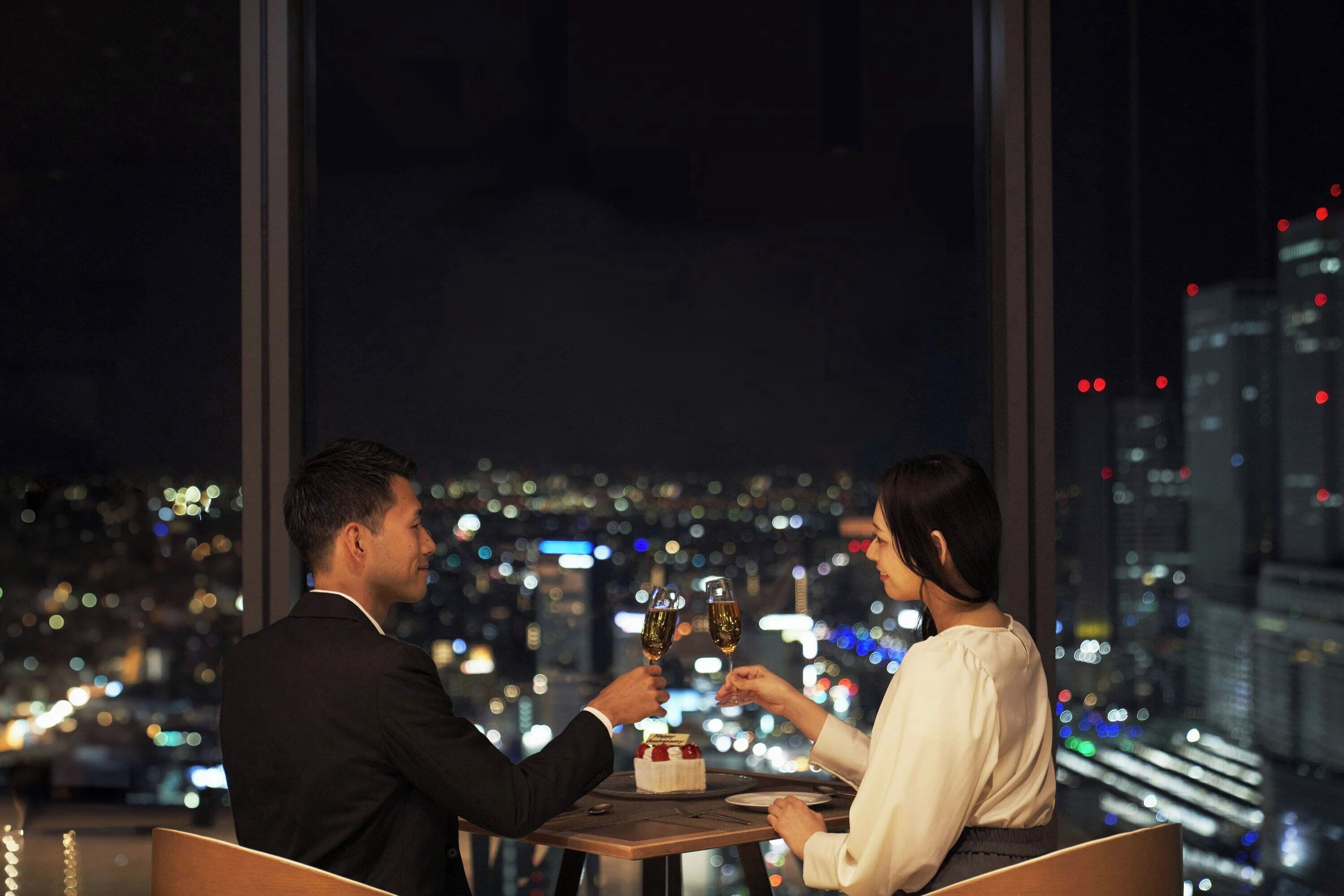 A couple toasting with champagne glasses at a table with a dessert, overlooking a cityscape at night.