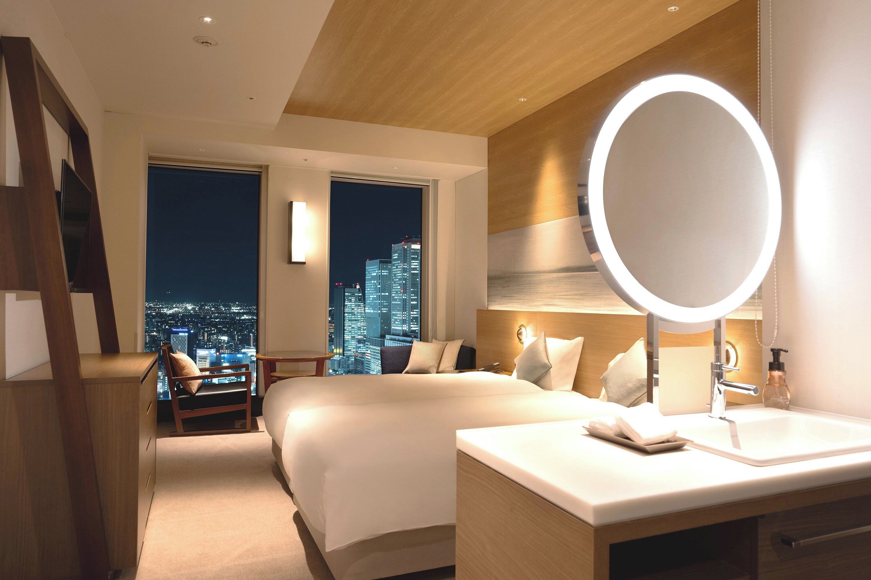 Habitación de hotel moderna con una cama grande, un espejo junto a la cama y vistas de la ciudad de noche.
